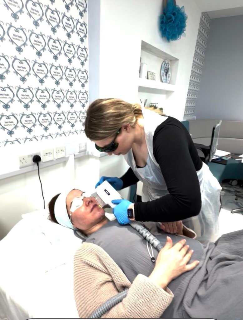 Practitioner treating a client’s face with a light-based acne therapy handpiece, wearing laser eyewear, gloves and an apron, with the client’s eyes shielded